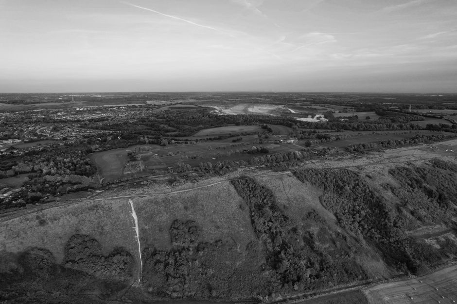 Aerial black and white photograph showcasing a rural landscape with rolling fields, patches of woodland, and scattered houses under a partly cloudy sky. The scenery includes winding roads and visible 