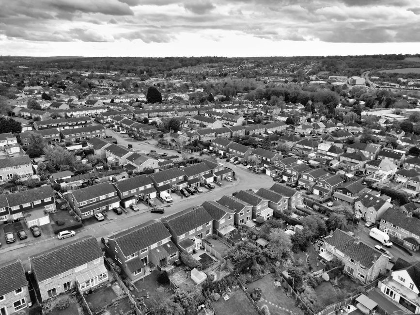 An aerial view in black and white showing a residential neighbourhood with rows of terraced and semi-detached houses, each with small front gardens, driveways, and parked cars along the streets. The h