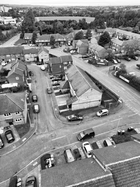 An aerial view in black and white showing a residential neighbourhood with rows of terraced and semi-detached houses, each with small front gardens, driveways, and parked cars along the streets. The h