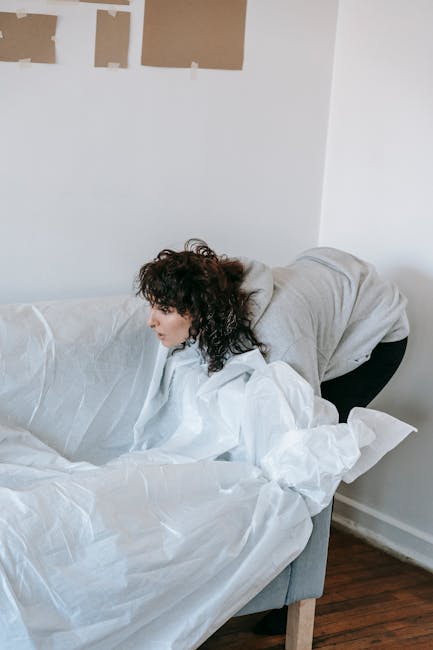 A woman with curly dark hair, dressed in a light grey hoodie and dark trousers, is leaning over a white sofa inside a room with white walls. She is handling protective packing materials such as white tissue or moving blankets, which are draped over the sofa to safeguard furniture during a house move. In the background, on the white wall, there are three brown paper templates or paint swatches taped up, suggesting ongoing interior work or preparation for a relocation. The floor is made of dark wood, and natural light illuminates the scene, highlighting the detailed textures of the fabric, packing materials, and the woman's focused posture. The image captures a moment of packing or furniture protection in the context of house removals, consistent with the services offered by Man with Van Biggin Hill, whose expertise includes home relocation and furniture transport preparations.