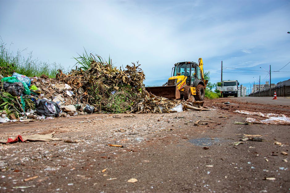 A yellow backhoe loader with a large front bucket is positioned on a paved road, actively clearing and scooping up a pile of mixed waste, including cardboard, plastic bags, and plant debris, near a grassy area. The scene is outdoors under a partly cloudy sky, with the machine's operator visible inside the cab. In the background, there is a white van parked on the side of the road, alongside some temporary traffic cones, indicating ongoing waste removal or clearance work. To the right, there are power lines and a building, suggesting an urban or suburban environment. The detailed image captures the process of waste collection and the use of heavy machinery typical in home relocation or large-scale house removals, conforming to the context of managing bulky waste rules for movers in the London Borough of Bromley, as outlined by Man with Van Biggin Hill.