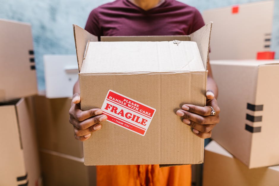A person holding an open cardboard box with a red and white 'Fragile Handle with Care' sticker, standing amidst several other packed boxes in a room, likely part of a home relocation or moving process. The individual’s hands, with a silver ring visible on one finger, grasp the box firmly, revealing the interior which appears empty. The background includes additional cardboard boxes stacked on the floor and against the wall, some sealed with black tape, indicating an organized packing setup. The environment is well-lit, suggesting indoor natural or artificial lighting, and the boxes are made of brown corrugated cardboard, used for packing household items for a move. These details reflect a professional moving or packing service, such as those offered by Man with Van Biggin Hill, involved in furniture transport and relocation logistics, consistent with guidelines for house removals and in accordance with London Borough of Bromley bulky waste rules.