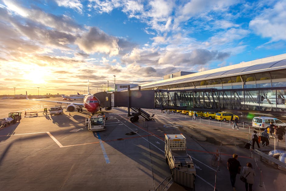 A view of an airport terminal at sunset showing a red and white airplane parked at the gate with a black passenger jet bridge extending from the building to the aircraft door. The tarmac features several ground crew members in high-visibility vests, alongside luggage carts, trolleys, and service vehicles. The airport building has large glass windows reflecting the sky, which is filled with scattered clouds illuminated by the setting sun. The scene includes a paved area with marked parking spaces and a few yellow taxis and cars parked nearby. The overall environment is bright with natural light, indicating the late afternoon or early evening, and captures the typical busy atmosphere of an airport ready for departing or arriving flights. Man with Van Biggin Hill, specializing in removals and home relocations, could utilize similar scenes to illustrate efficient packing and transport logistics involved in house removals near an airport.