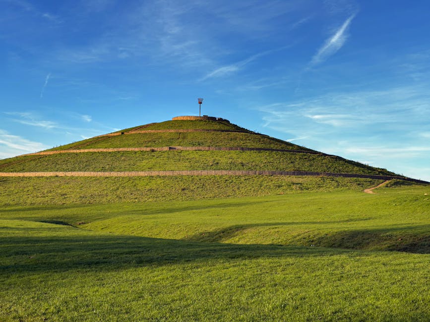 In the image, a large, grass-covered hill with several paved, terraced pathways climbing up its incline is visible. At the top of the hill, there is a small, white, lattice-style observation tower or weather station structure. The foreground shows a wide, green grassy area with gentle undulations, under a clear blue sky with a few wispy clouds. The scene is well-lit with natural sunlight, highlighting the vibrant green of the grass and the crispness of the blue sky. This outdoor landscape may be associated with publicly accessible areas near Biggin Hill, potentially relevant for house removals or relocation services, such as providing suitable access routes for moving vehicles or viewing points for planning home relocations, as highlighted on the page about moving near Biggin Hill Airport with access and parking tips.