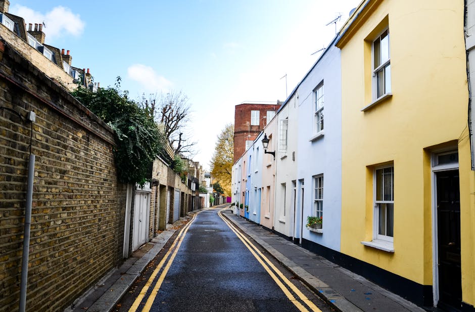 A narrow residential street in Biggin Hill showing a wet pavement with double yellow lines along the curb. On the right side, there are colorful terraced houses painted in pastel shades of yellow, white, and light blue, with white-framed windows and small flower pots on the sills. Some houses have external wall-mounted lamps. On the left side, a weathered brick wall separates the street from a row of garages and garden areas, with greenery creeping over parts of the wall. A large red brick building is visible further down the street amidst leafless trees, indicating autumn or early winter. The sky is partly cloudy with patches of blue, and soft daylight illuminates the scene. This setting reflects a typical small-street environment suitable for house removals, where furniture and packing materials are likely being prepared for transport by professionals like Man with Van Biggin Hill, as part of a home relocation or moving process.