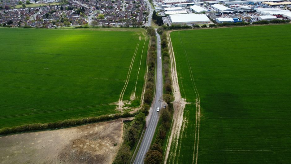 Aerial view of a rural area showing a narrow lane road running between two large green fields with visible crop lines, bordered by hedges and trees. The road has a few vehicles, including a white van, which is likely involved in a home relocation or furniture transport process. In the background, there are industrial and commercial buildings, indicating proximity to a developed area. The scene is captured during daylight, with clear weather and natural lighting. This setting reflects the logistical aspects of move planning and access for small-street teams, as provided by Man with Van Biggin Hill, focusing on moving services within tight residential access points.