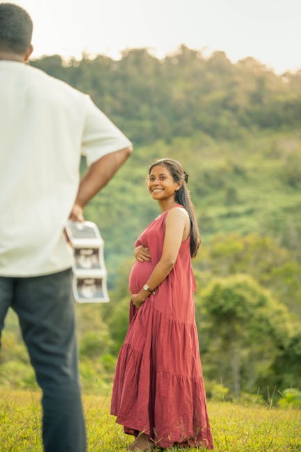 A young pregnant woman outdoors on a grassy field, smiling and looking at a man holding a clipboard, with trees and a hillside in the background. She is wearing a long, sleeveless, red dress and has her arms crossed over her belly. The man, seen from behind, is dressed in a light-colored shirt and dark trousers. The scene is brightly lit by natural sunlight, creating a warm and cheerful atmosphere, which could be part of a casual photo shoot or personal photo session. This image does not relate directly to house removals or moving services but captures a moment of happiness during an outdoor activity.
