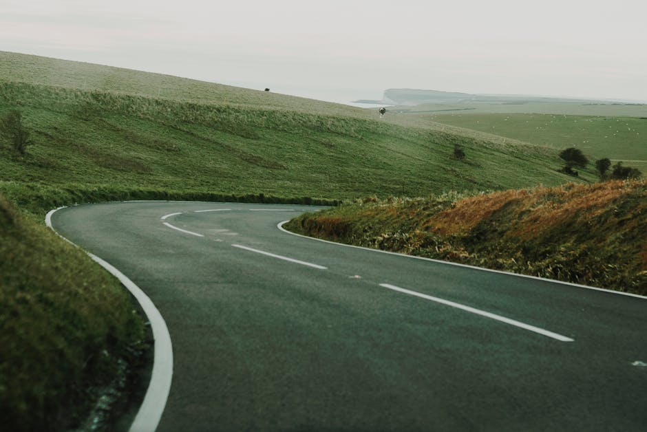 A winding rural road with smooth asphalt surface and white dashed lane markings curves through open countryside with gently rolling green hills and sparse trees. The road appears to be in a quiet, peaceful area with no vehicles or pedestrians visible, and the landscape extends into the distance under a cloudy, overcast sky. This scene provides a tranquil backdrop for discussions about home relocation or furniture transport logistics, as featured in the page titled 'Station Road to Church Road moves in Biggin Hill' by Man with Van Biggin Hill, specialists in removals. The natural lighting highlights the lush greenery and the curvature of the road, emphasizing the importance of careful planning in moving and transportation processes across rural areas.